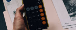 a hand holding a phone over a stack of papers and a pen